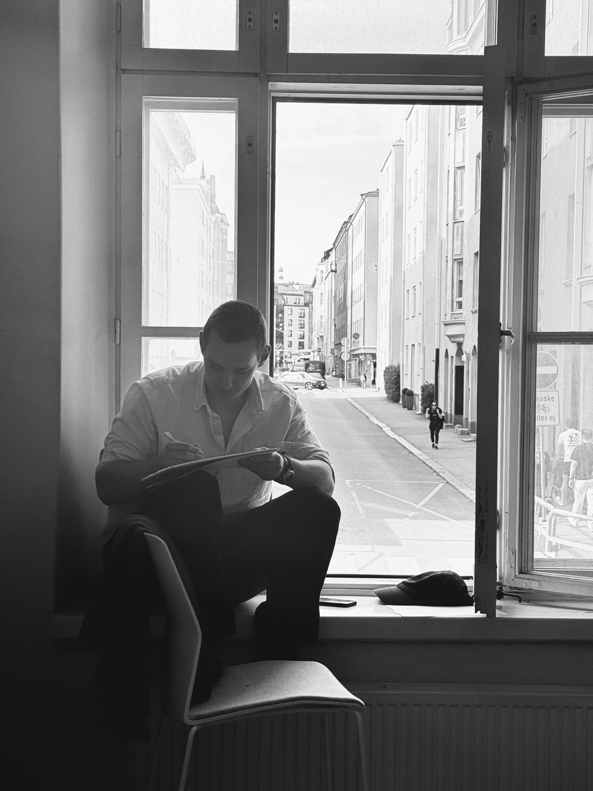 Black-and-white photo of Petr sitting on a window sill, writing in a notebook, with a city street outside the open window